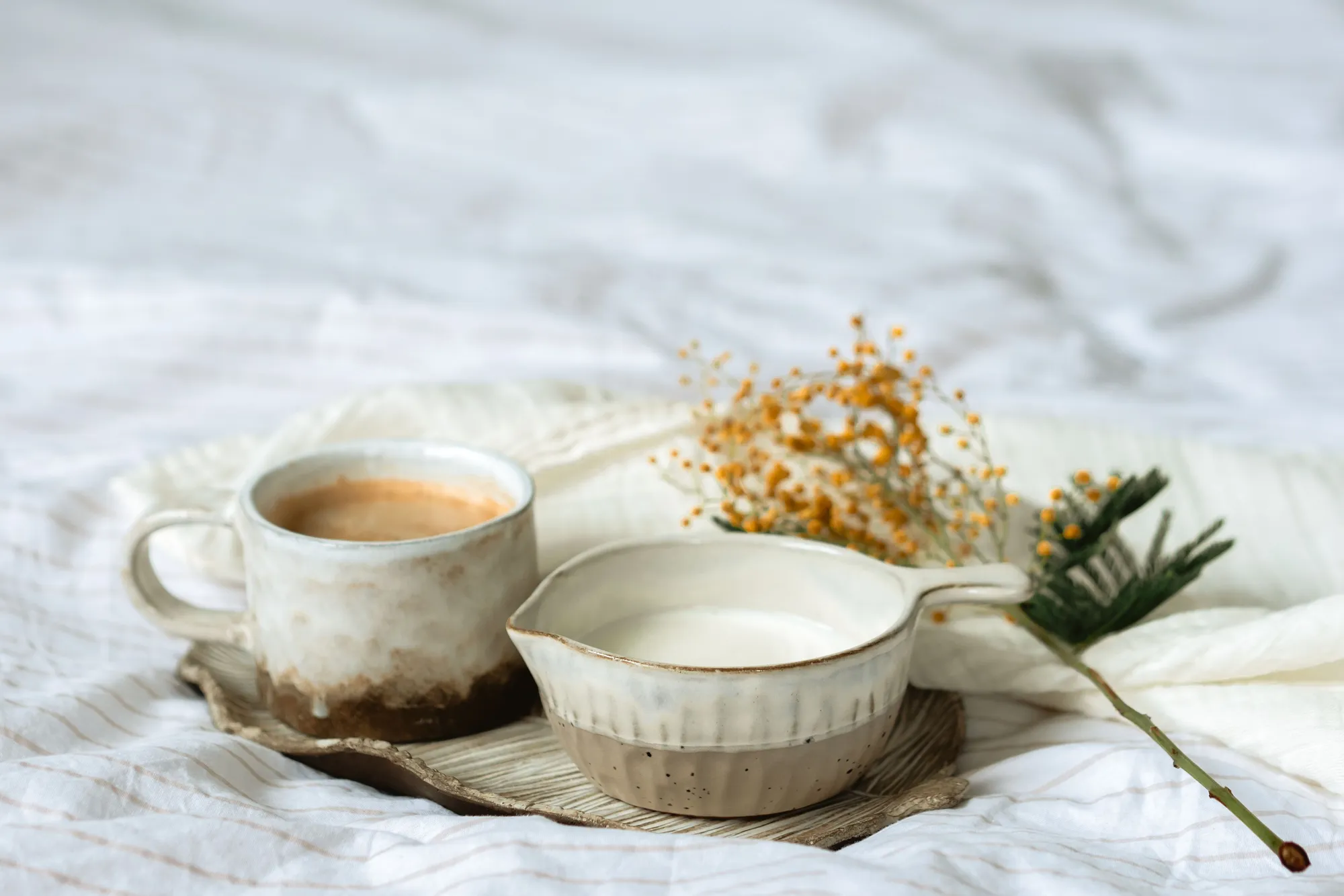 Two handmade ceramic cups, one holding coffee, on a ceramic tray with a sprig of dried yellow flowers on soft white linen
