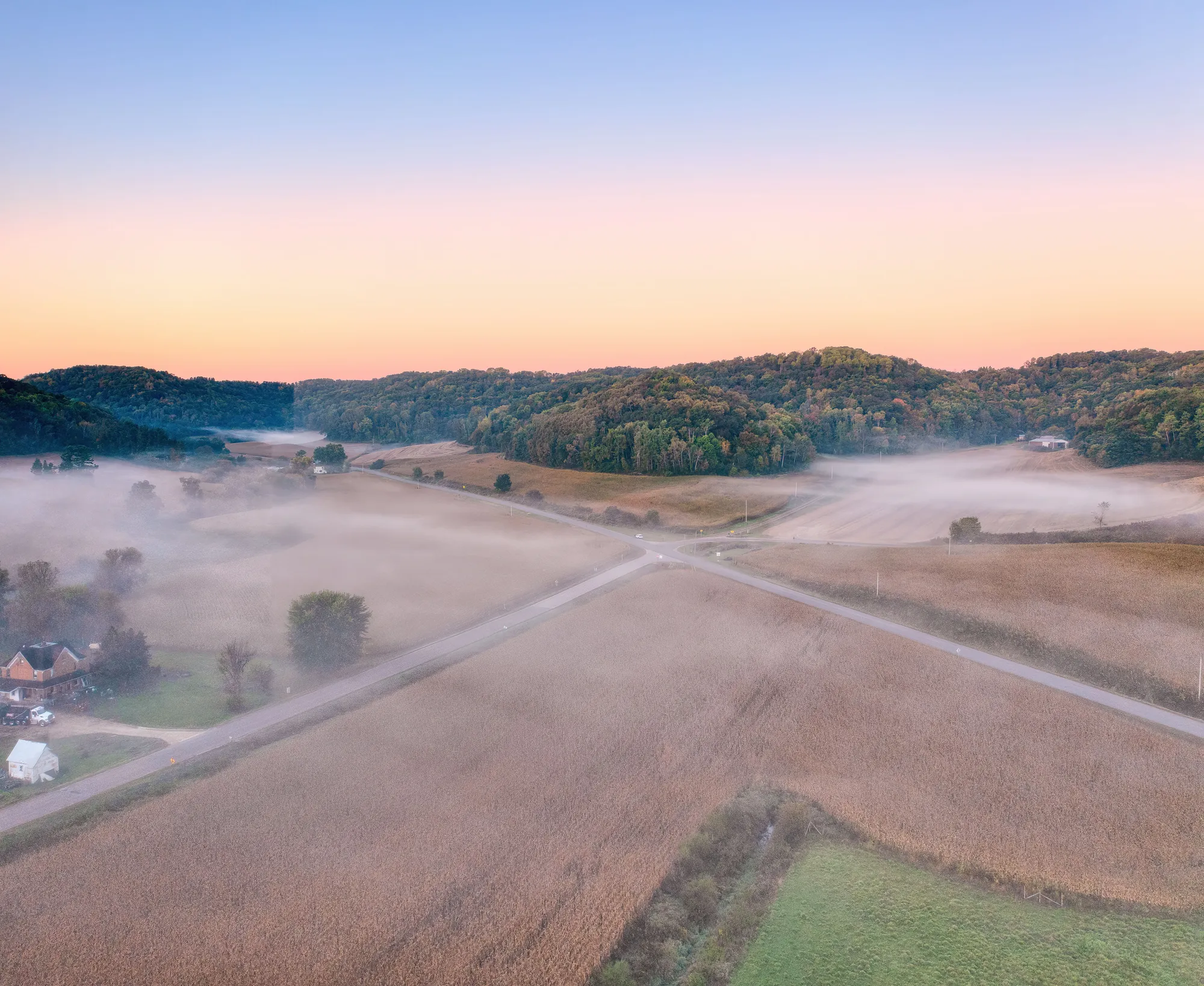 Aerial view of two quiet country roads crossing through misty farmland and wooded hills at sunrise