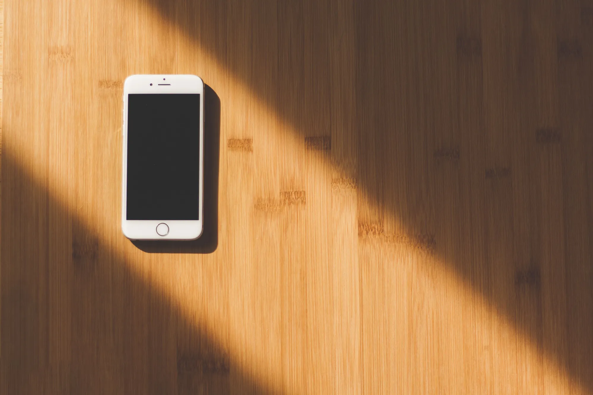 A smartphone with a dark screen lying on a wooden table in a diagonal band of warm sunlight